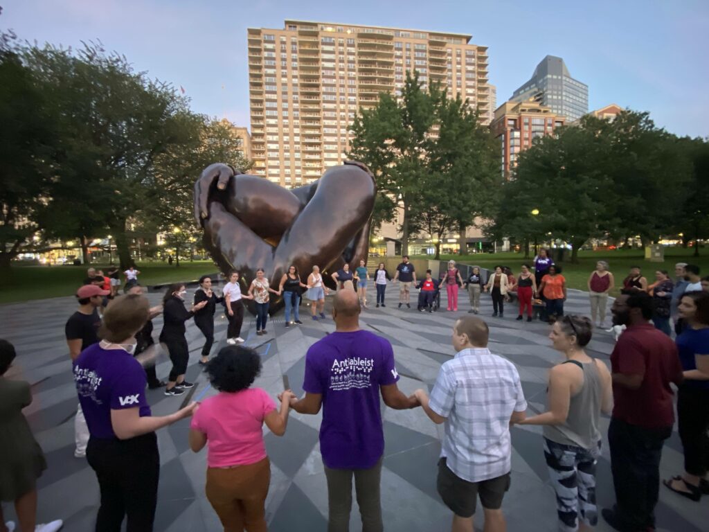 A diverse community of various colors, disabled and non-disabled holds hands in a circle in front of the majestic embrace memorial, a bronze hug.