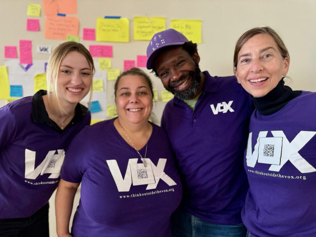 Vox team members standing and smiling with arms around each other wearing purple Vox shirts, jeans and dark shoes in front of a wall decorated with a paper map for a 2D organizational activity. From left to right: Ingrid O’Dell, a tall white woman with blonde hair, blue eyes and freckles has her hair tied back; Sam Gould, an Ashkenazi Jewish woman with salt and pepper hair in a bun; Christopher Robinson, an African-American man with black hair, medium locs, and a beard wears a purple Vox hat with his head tilted down and to the side; Kristin Johnson, a Deaf white woman with light brown shoulder length hair tied back and holding a pair of clear glasses at her hip.
