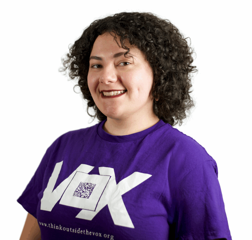 Olivia, a Hard of Hearing mixed Latinx with olive skin and dark brown curly hair wears a purple Vox shirt and cranberry lipstick, smiling