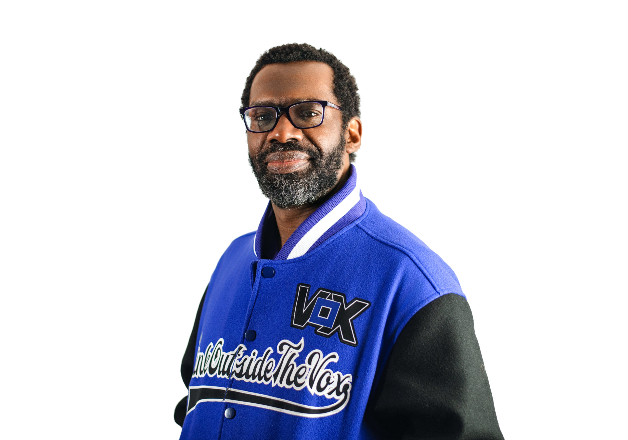 an African-American man with black hair, medium locs, glasses and a seasonal beard wears a purple Vox letterman jacket with the Vox logo, and Think Outside The Vox black text across the front