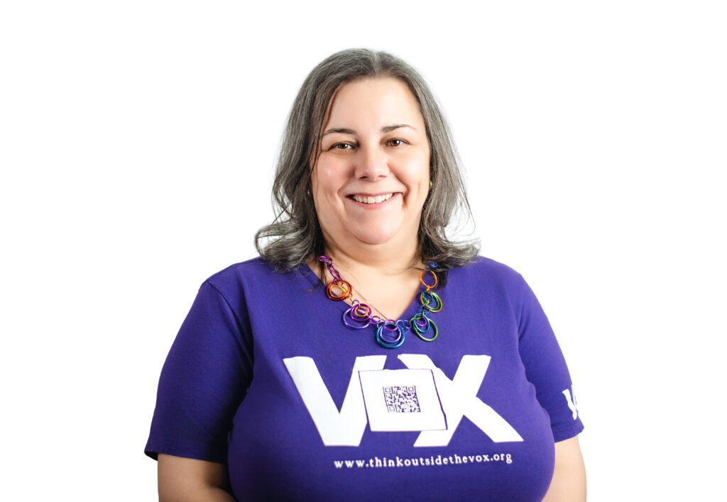 An Askenazi Jewish woman with salt and pepper hair wearing a Purple Vox t shirt with a QR code. She wears a colorful chunky chain necklace and smiles at the camera. She has a double chin and dimples.