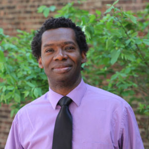 Photo of Chris Robinson, African American man in a purple shirt, standing outdoors in front of a green leafy backdrop, smiling
