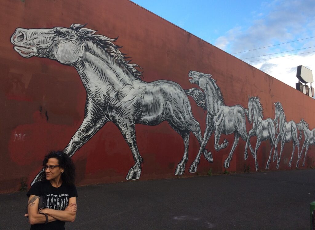 An enormous mural of running horses in black and white on a red wall. Cheryl, a white Ashkenazi woman with olive skin and long, curly brown hair, stands with arms folded, her forearms chiseled from too many years typing, staring off in the direction the horses are headed. She has a solemn crow tattooed on one arm and wears a black t-shirt with white text, “No More Spoons” and a row of knives and daggers.