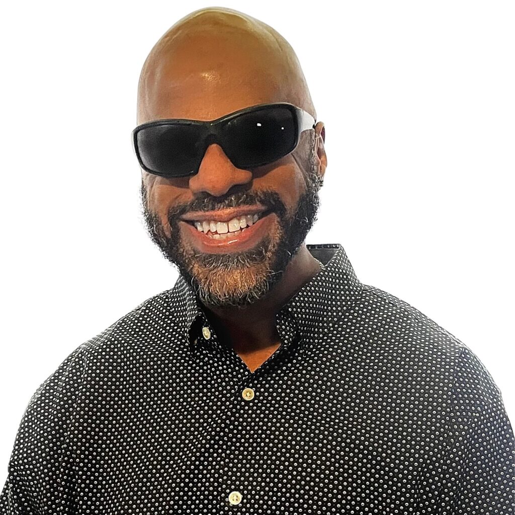 Thomas Reid, a brown skin Black man with a clean-shaven bald head and full neat beard,  smiles into the camera. He wears dark shades and a black and white checkered button up shirt.