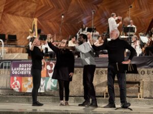 the ASL team performing in front of the Boston Landmarks Orchestra at the Hatch Shell. All stand with expressions of hope and promise, all gesturing something different with one hand.