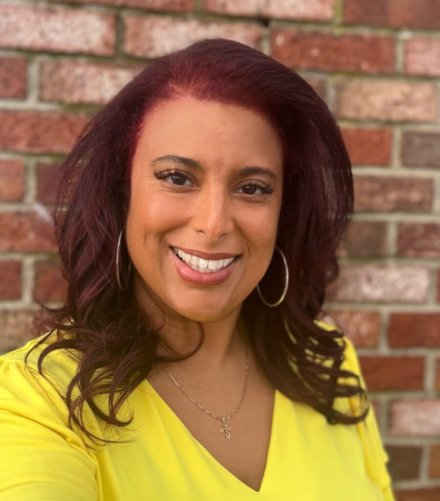 An Afro Indigenous woman with auburn hair, gold hoop earrings and a bright yellow top smiles at the camera