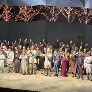 Entire cast of actors and singers for Macbeth on stage in their white and neutral colored utilitarian and traditional military regalia bowing, waving and smiling. In the center front row, Léon Jerfita, a white male with short silver hair, a short white beard and one blue and brown eye signs “I Love you” with both hands towards the audience next to Director Steve Mahler, a tall white man in a blue suit.