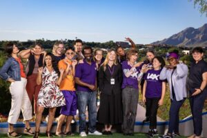large group photo of Massachusetts-based arts professionals (former and current) standing, posing and smiling in front of an Arizona landscape with blue skies, a mountain on the right, cacti and desert plants and trees. Some pose with hands under chin, a peace sign, pointing and laughing. From left to right: from RAW Art Works, Laura Smith-Centeno, Rosario Ubiera-Minaya, Mariah Neuroth. Formerly at WBUR and now in Texas at Report for America, Cristela Guerra. Arts Connect International mica rose. Double Edge Theatre Travis Coe. Think Outside the Vox Christopher and Aimee Robinson, Ingrid O’Dell, Maria Hendricks, Kristin Johnson, Sam Gould, Olivia Reinebach, and ASL/English interpreters Shonnese Devon Grant and Rae Heller.